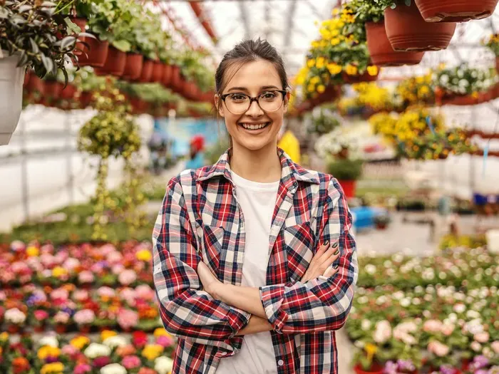 Young woman wearing glasses and plaid shirt smiling in a colorful greenhouse filled with hanging and potted flowers